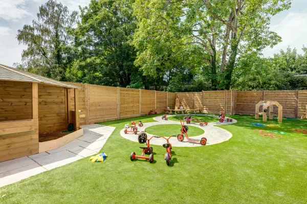 Red-and-black tricycles and ride-on toys are scattered along a painted circular path on artificial turf, in a fenced backyard playground with a wooden playhouse, archway, and tall trees.