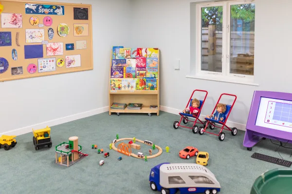 Toy vehicles and a wooden train set — scattered on a carpeted playroom floor; two dolls sit in red strollers near a bookshelf, purple touchscreen, window, and a bulletin board labeled "Space".