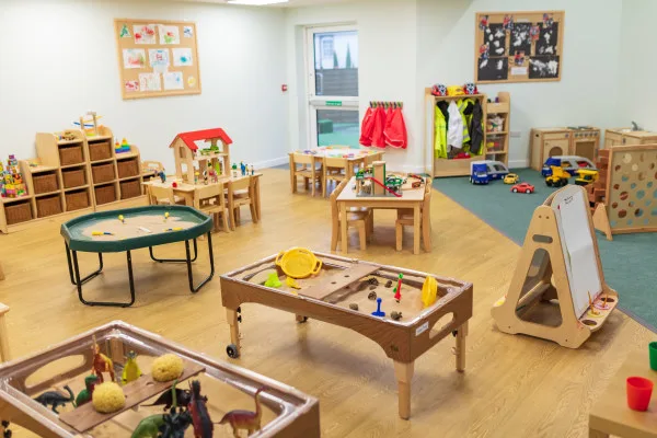 Wooden activity tables hold sand, toy animals, and manipulatives; small tables and chairs sit nearby, with cubbies, coat hooks, and toy cars arranged across a well-lit preschool classroom.