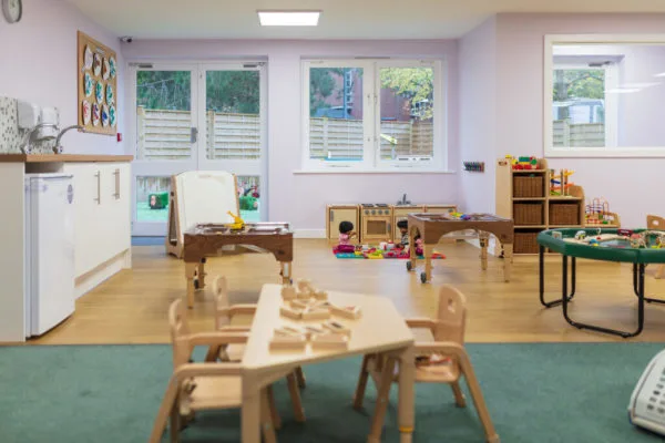 Small wooden table holds scattered wooden shapes; surrounding chairs face it in a bright nursery playroom with carpeted area, toy kitchen, activity tables, shelves, and windows showing a yard.