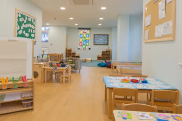 A children's classroom: small wooden tables and chairs hold craft supplies and toys, arranged across a bright, pastel-painted daycare room with shelves, bulletin boards, cushions, and a play area.