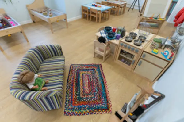 Small striped armchair holds a seated doll reading a book, on a colorful braided rug, surrounded by a wooden play kitchen, low tables and toy stations in a bright children's playroom.