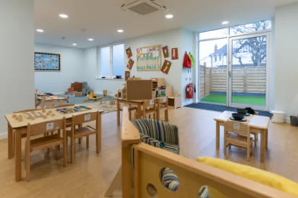 Child-sized wooden tables and chairs arranged for play, holding toys and materials, inside a bright preschool classroom with displays on walls and a sliding glass door to an outdoor area.