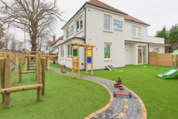 Two-story white daycare building overlooks a fenced playground; a toy tricycle sits on a curved paved path amid artificial turf, climbing equipment, and slides. Text on building: "Perfect Start Day Nursery".
