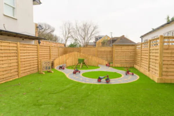 Ride-on tricycles parked along a painted circular track, arranged around a small activity table in a grassy, fenced backyard play area with neighboring houses.