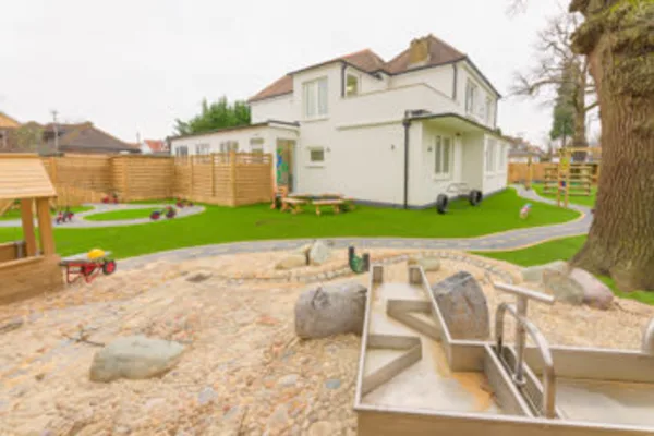 Metal water channels sit atop a sandy, stony play area in front of a white two-story building, surrounded by artificial grass, wooden fencing, picnic benches, pathways, and a large tree.