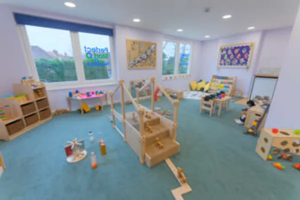 A wooden climbing play structure stands center, surrounded by toys, small tables and cushions in a bright, carpeted preschool classroom; window sign reads "Perfect Start".