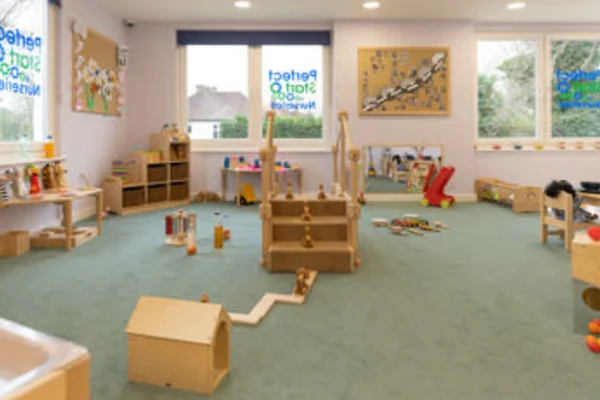 Wooden toy structures and blocks arranged for play on a green-carpeted nursery floor, with low shelves and colorful toys around; windows bear text: "Perfect Start for Our Nursery".