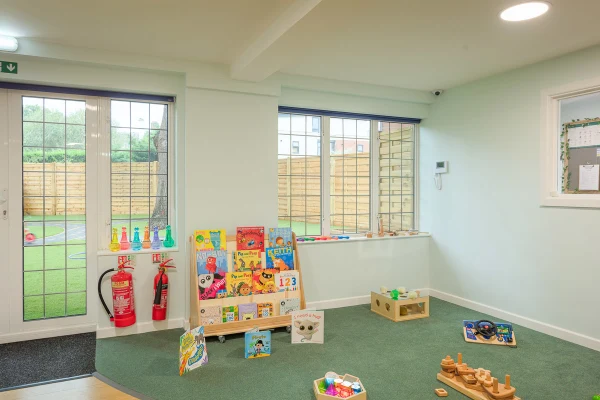 Low wooden bookshelf displays colorful children's books and toys on a green-carpeted play area by windows and a door with two fire extinguishers. Visible text: "123".