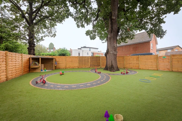 Mature tree anchors a fenced children's play area, surrounded by a circular paved track with scattered tricycles and hoops, set on artificial turf in a residential backyard.