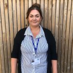 Woman standing, facing camera, smiling; wearing a black cardigan over a white shirt with black vertical stripes; blue 