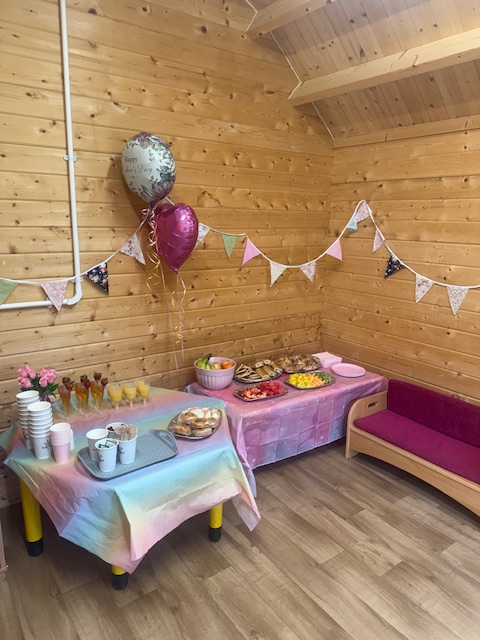 A cosy wooden room decorated with bunting, balloons, and pastel tablecloths. A food table is set with sandwiches, fruit, pastries, and drinks.