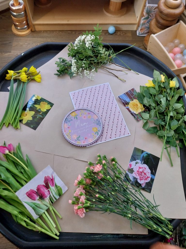 A creative activity table with fresh flowers, pictures of different flower types, and a ‘Mum’ plaque, inviting children to explore floral arrangements.