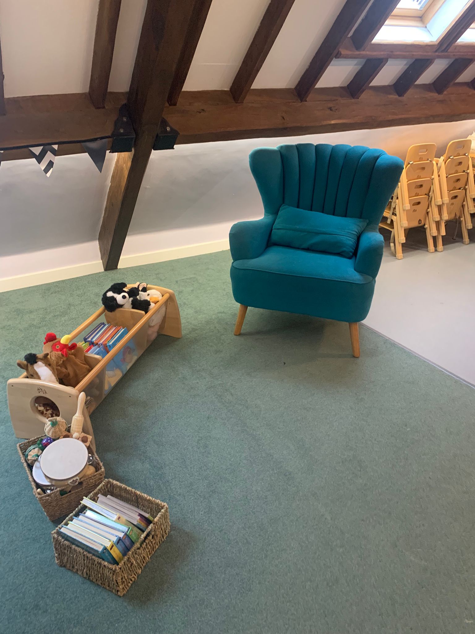 A teal upholstered armchair sits on a green carpet in an attic playroom, surrounded by wooden-beamed rafters, toy baskets and shelf, and a stack of small wooden children's chairs.