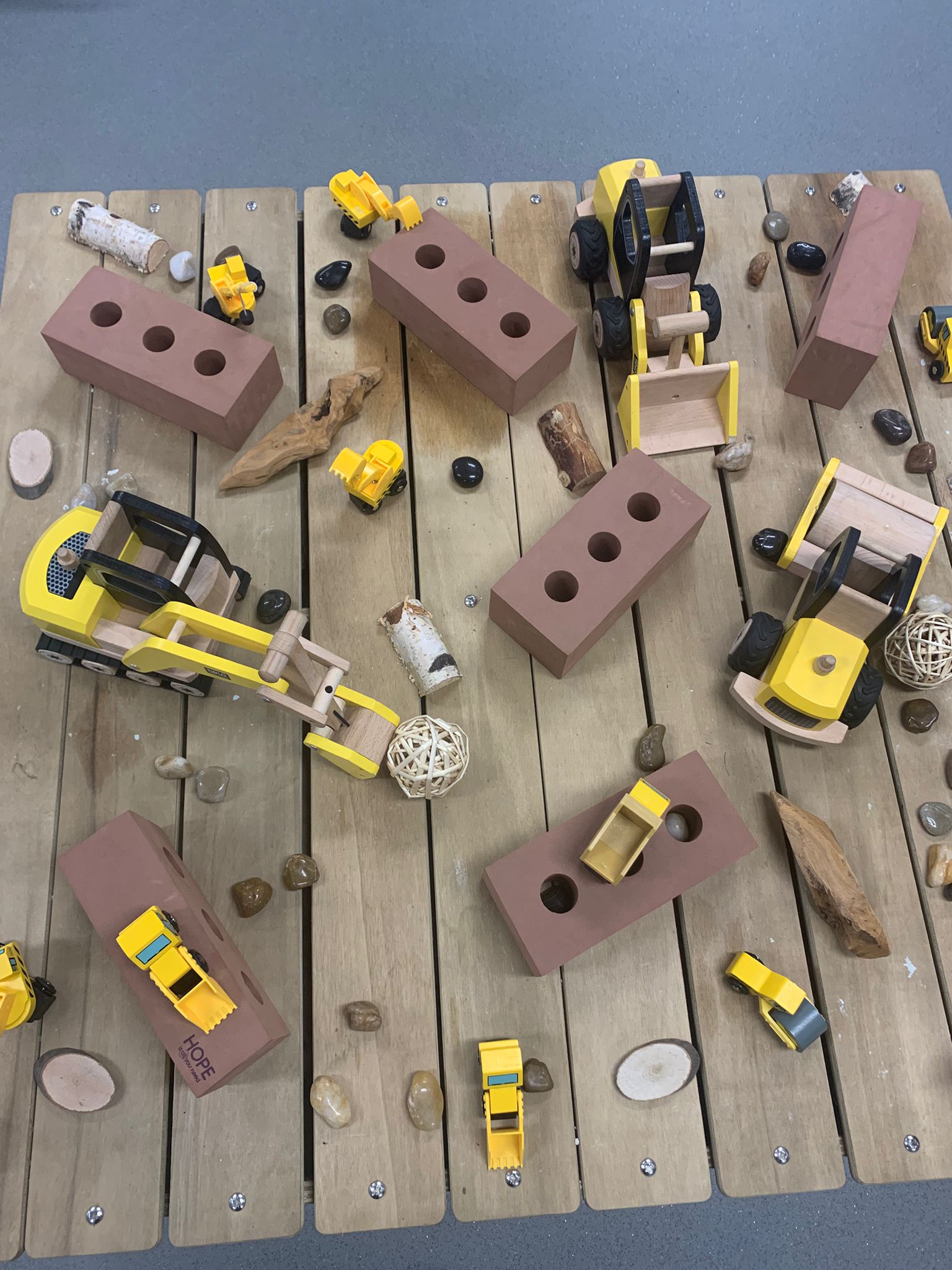 Several yellow toy construction vehicles move among brown three-holed toy bricks, scattered stones and wood pieces on a slatted wooden surface. Text on one brick: "HOPE".