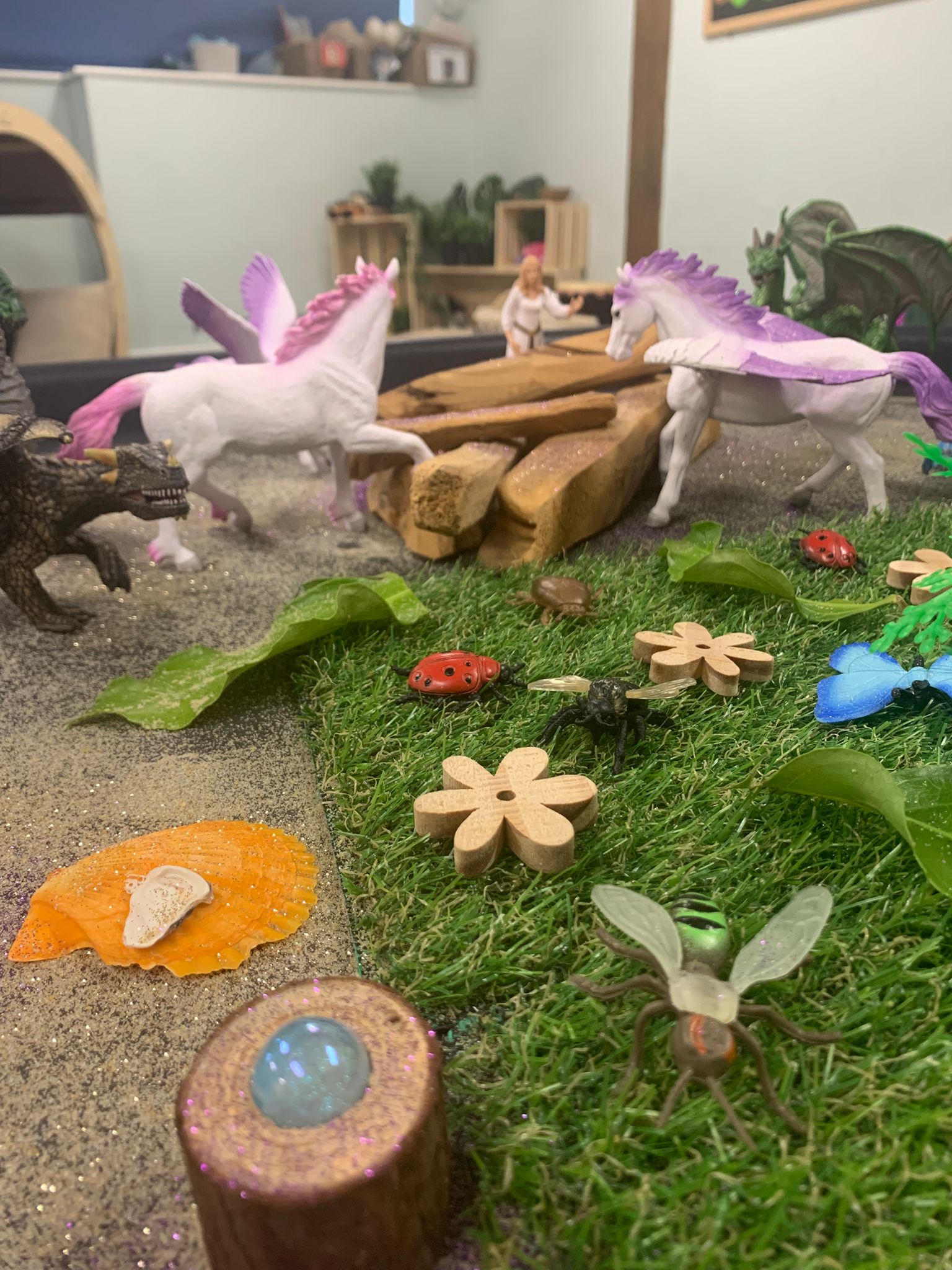 White plastic winged unicorns with purple manes approach each other across a wooden bridge in a tabletop play scene strewn with faux grass, wooden flowers, toy insects, shells and dragons.