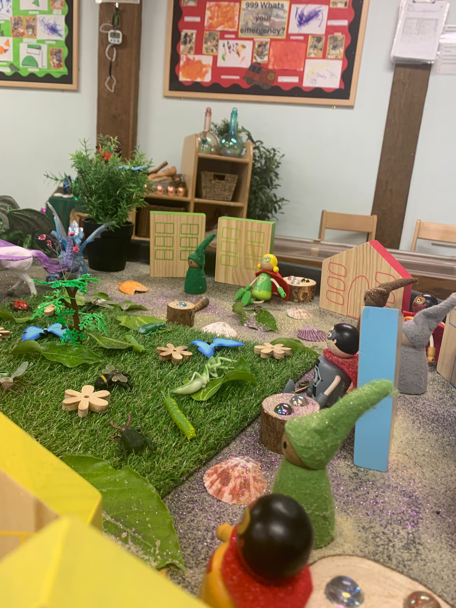 Wooden toy figures and gnome-like figurines gather around a miniature grassy play scene with wooden houses, shells, bugs, and craft items on a table in a classroom. Transcribed text: "999 What's your emergency?"