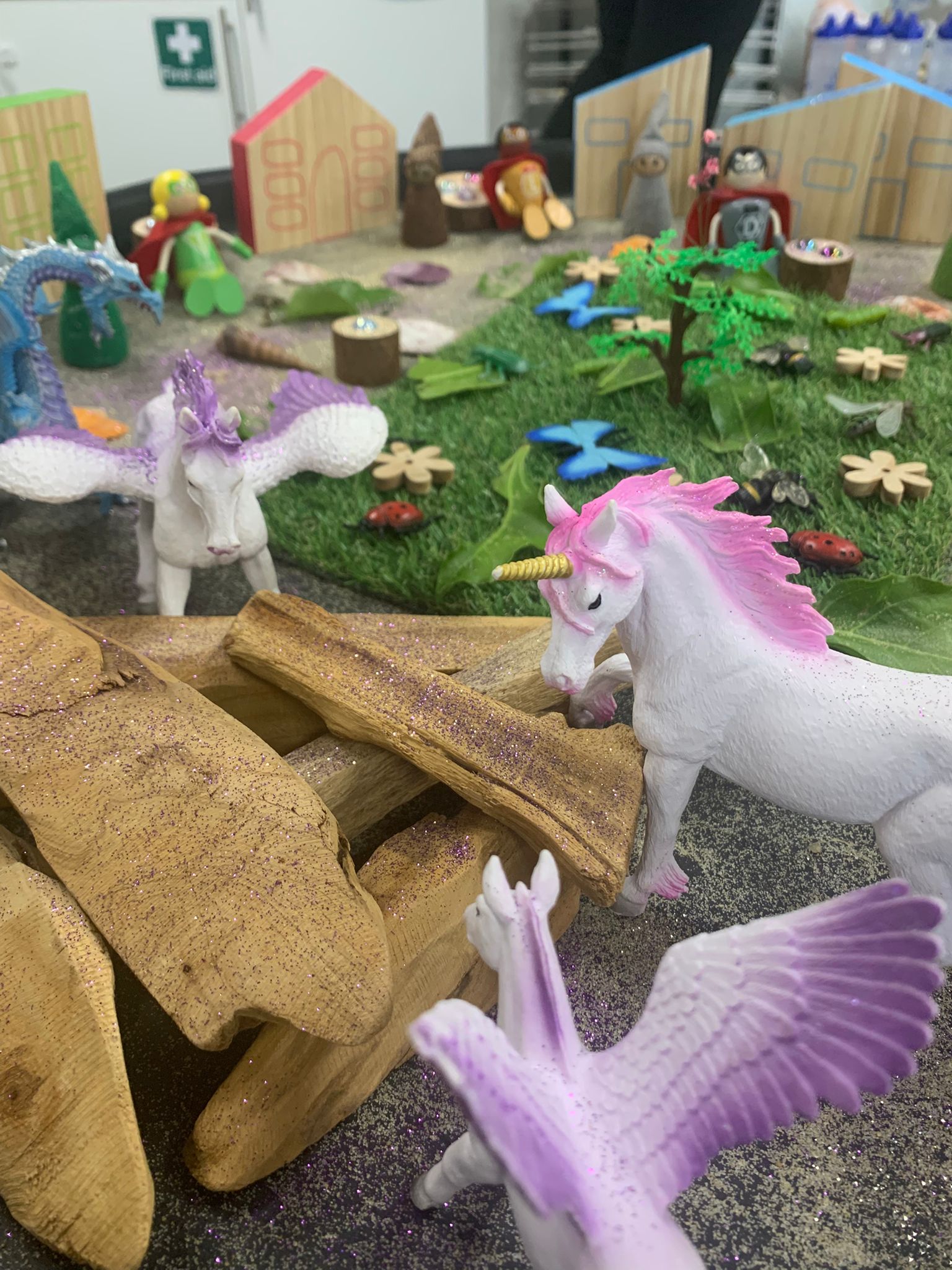 White unicorn figurine with pink mane and golden horn leans toward stacked driftwood pieces dusted with purple glitter; nearby winged ponies and miniature houses dot a grassy toy scene.