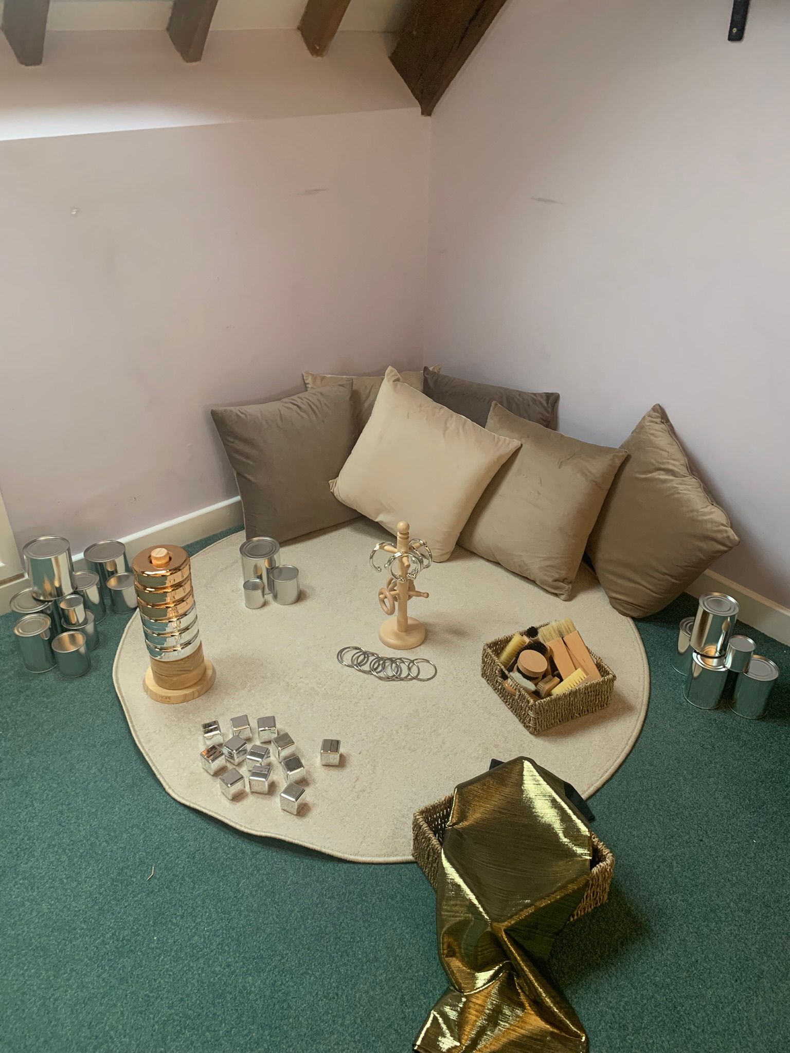 Round beige rug anchors corner seating area: neutral pillows against a pale wall, wooden jewelry stand and metallic stacking toys scattered on rug, baskets with brushes and gold fabric nearby.
