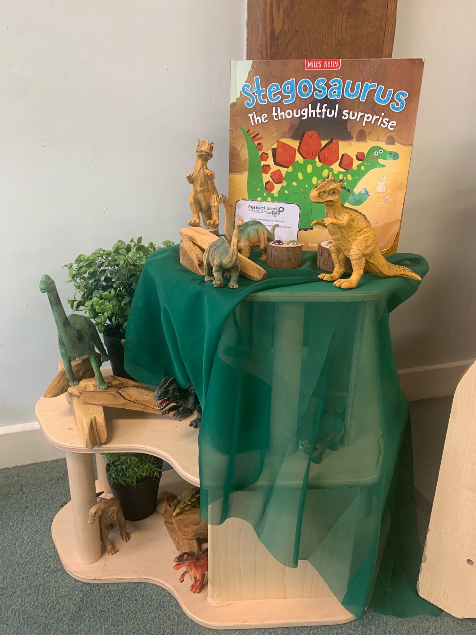 Children's book and plastic dinosaur figures arranged on a small wooden display draped with green fabric; toys stand among wooden blocks and potted plants in a carpeted corner.