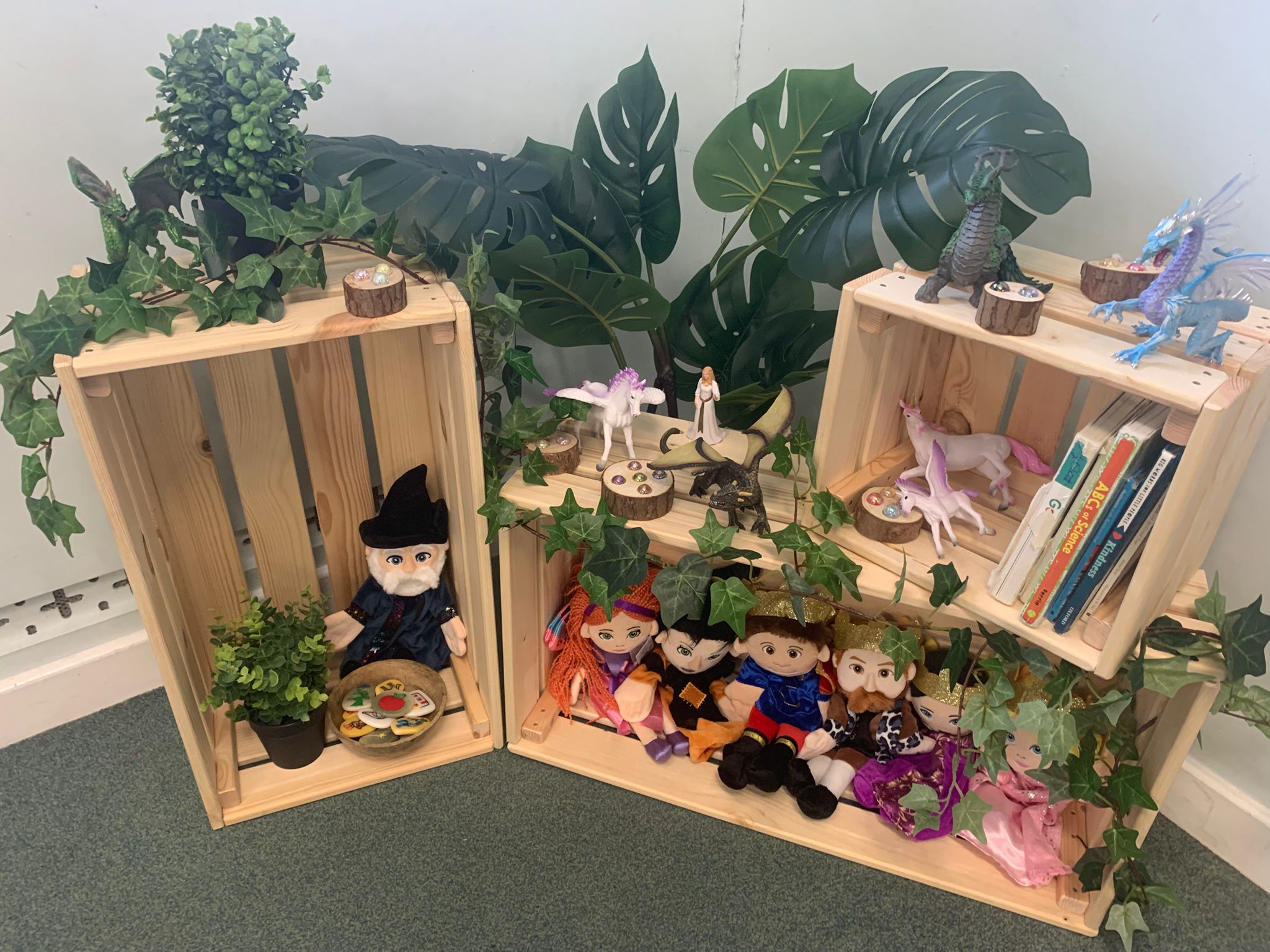 Wooden crates display plush puppets, toy dragons and unicorns arranged on shelves, accented with faux ivy, potted plants and wooden token dishes, stacked against a pale wall on a carpeted floor. No legible text.