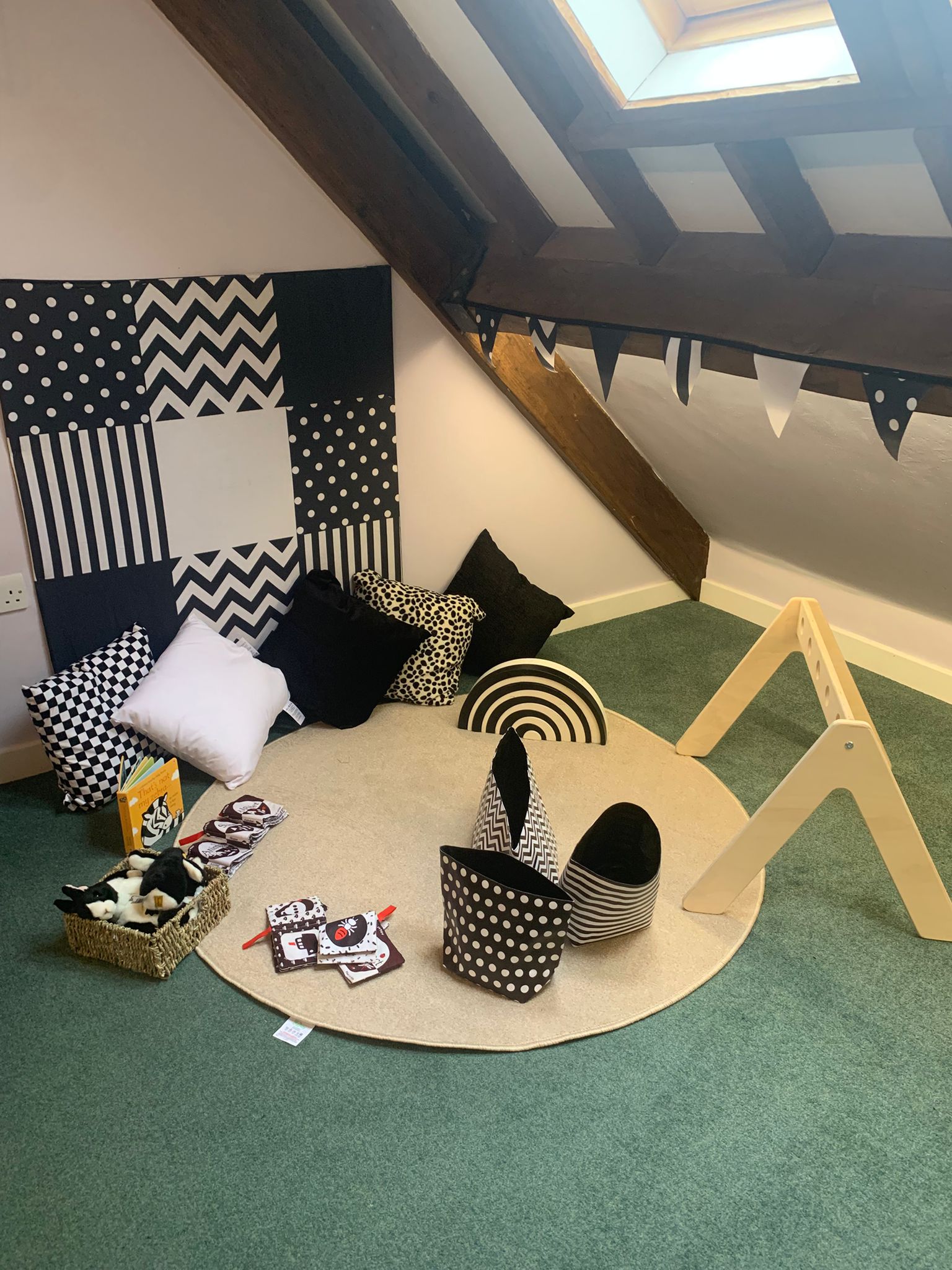 Round beige rug holds black-and-white soft toys, fabric baskets and cushions arranged for play; wooden climbing frame nearby in a sloped attic playroom beneath a skylight.