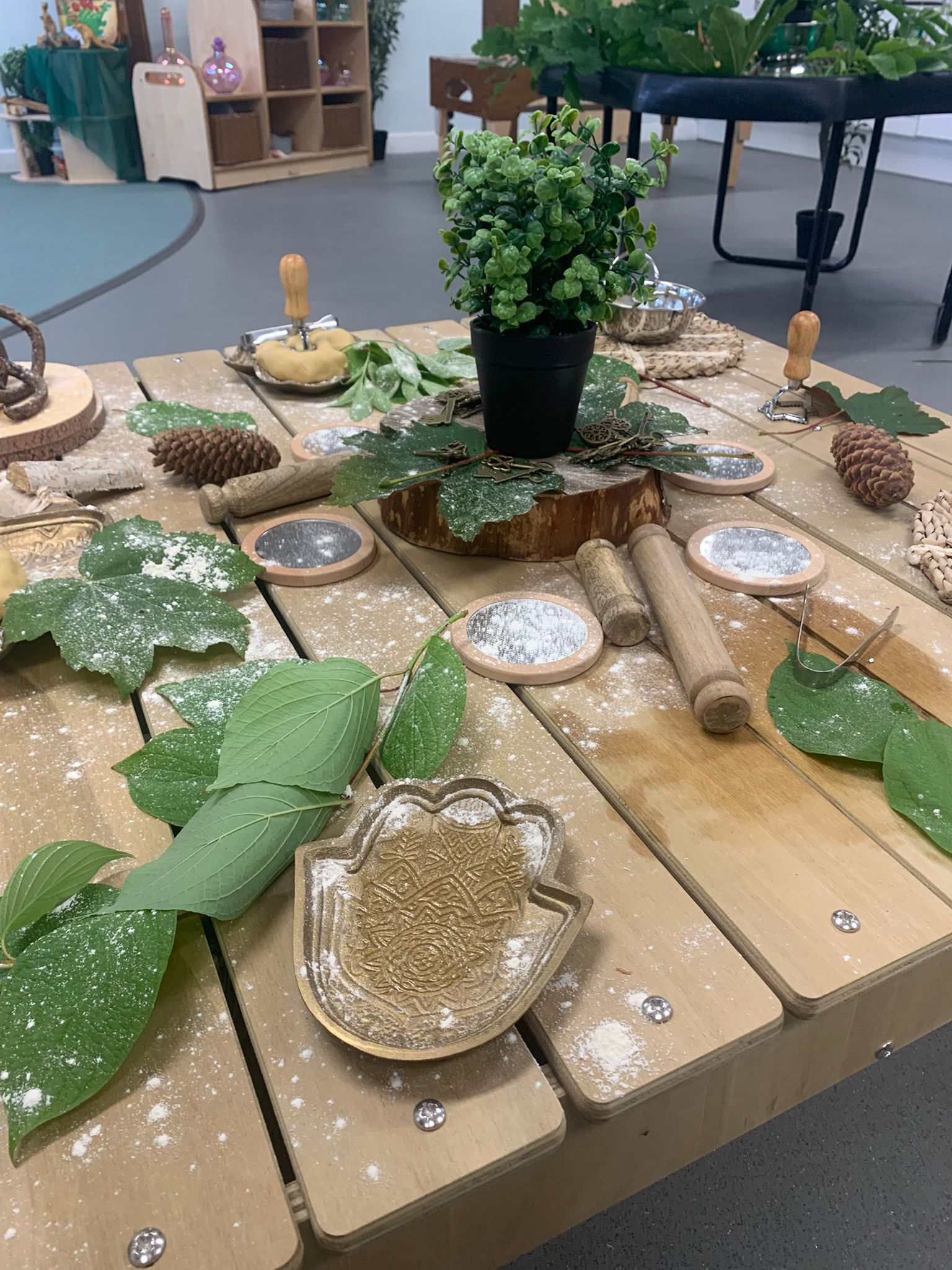 A small potted faux plant sits on a wooden slice, surrounded by leaves, pinecones, wooden rollers, decorative tray and flour-dusted craft tools on an indoor play-area wooden table.