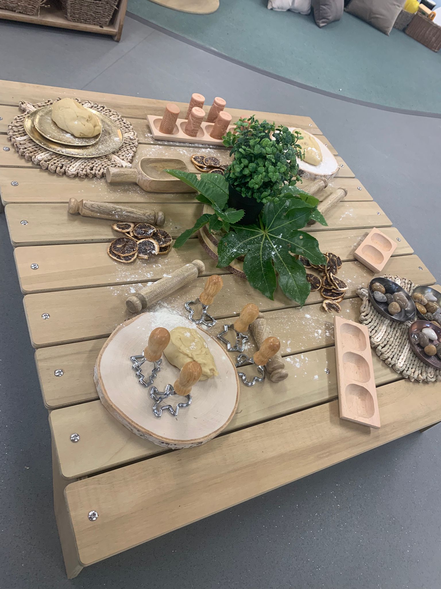 Low wooden table arranged with play dough, cookie cutters, rolling pins, dried orange slices and bowls, a potted plant centerpiece, flour dusted across an indoor classroom play area.