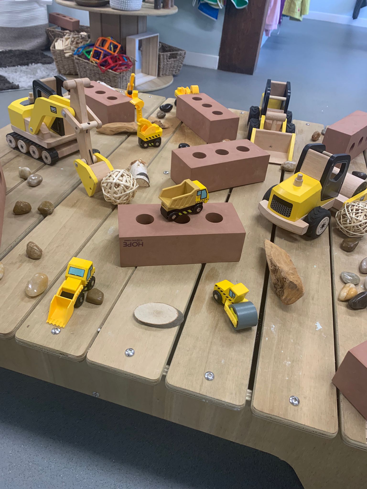 Wooden toy construction vehicles (excavator, dump truck, roller) sit scattered on toy bricks and a wooden activity table, surrounded by pebbles, wood pieces and wicker baskets in a playroom. Text on brick: "HOPE".