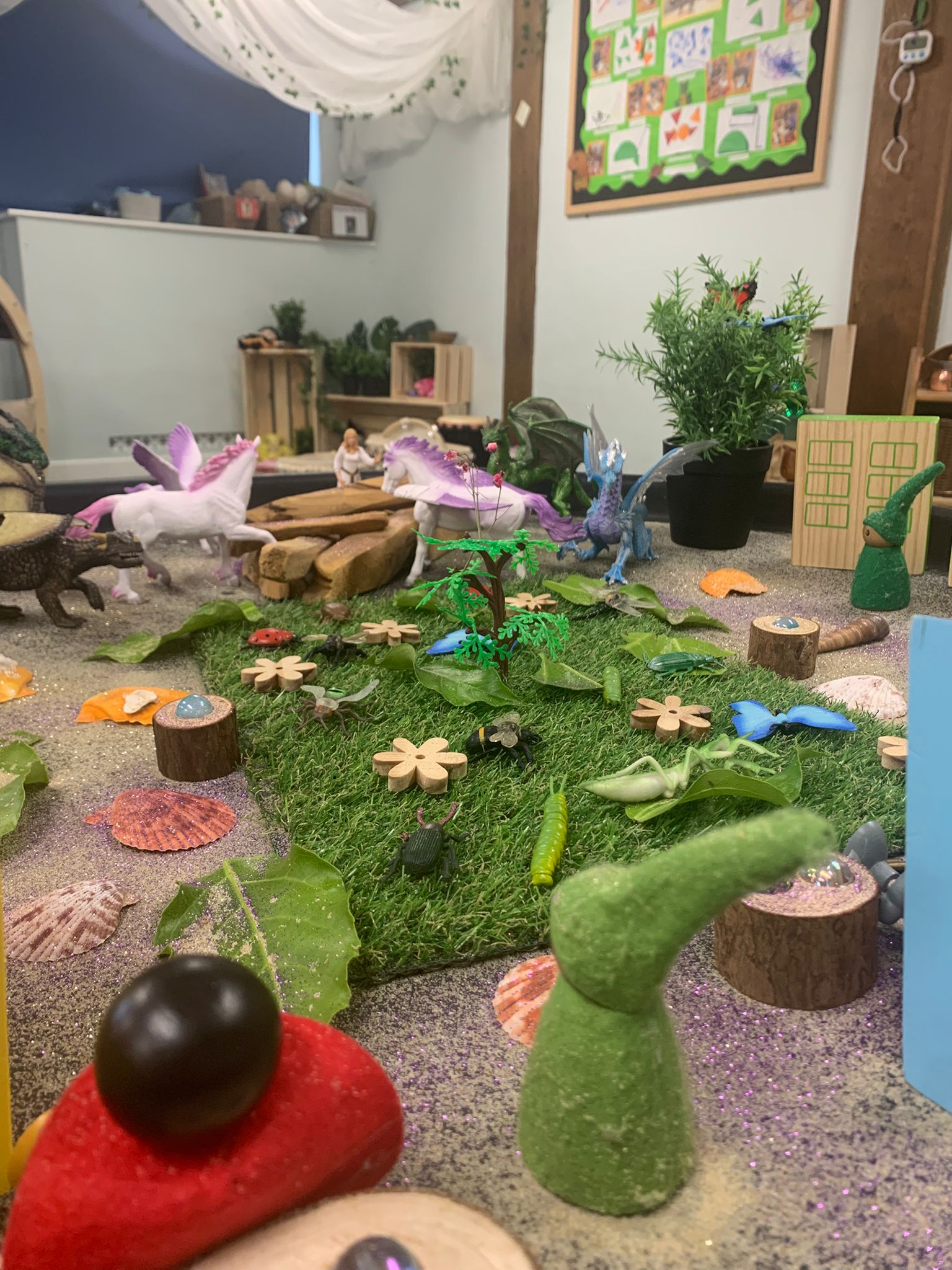 Toy fantasy scene: unicorns, a dragon, insects and plant figures arranged on fake grass and sand, creating a miniature landscape inside a classroom play area with shelves and bulletin board.