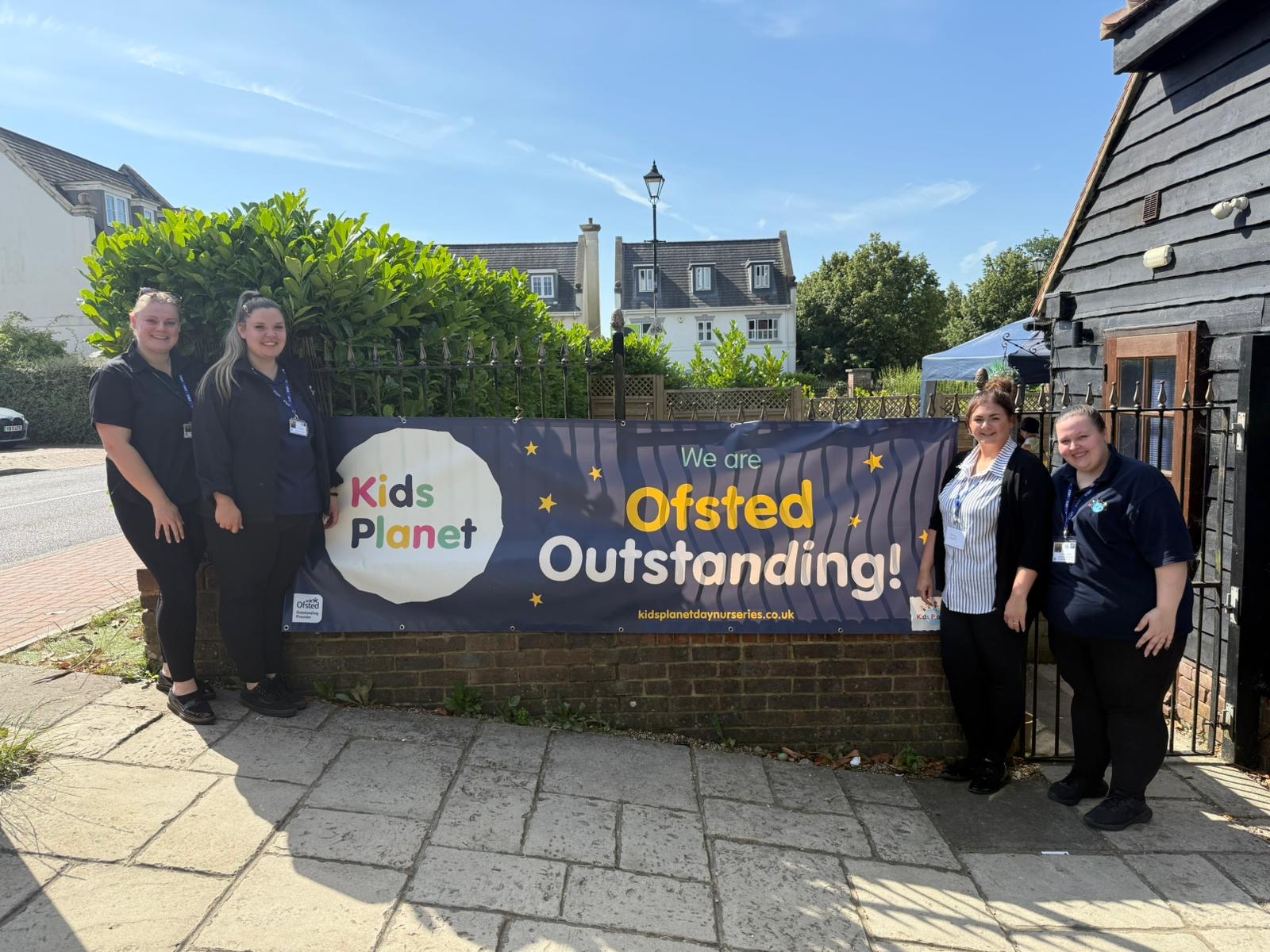 Banner reading "Kids Planet" and "We are Ofsted Outstanding!" displayed on a brick wall; four nursery staff stand smiling beside it on a sunny sidewalk in front of homes and a gated entrance.