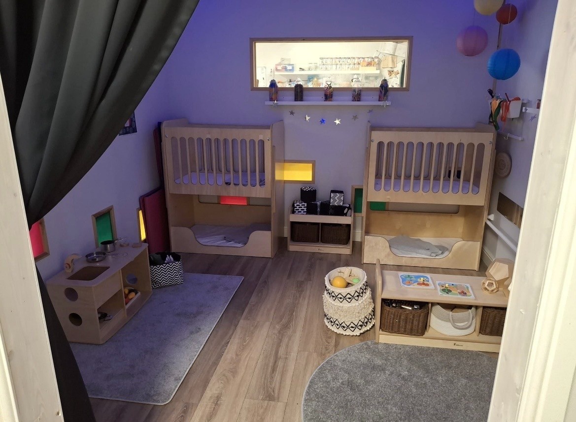 Two wooden toddler bunks sit empty, each with a lower nap cubby and guardrail; the cozy preschool playroom has soft rugs, low shelves with baskets, colorful inset windows, and hanging lanterns.