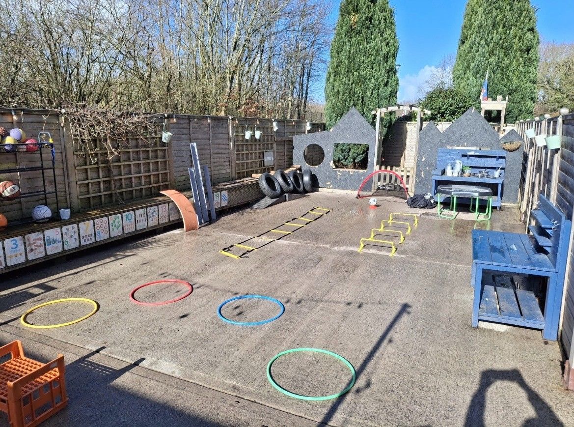 Outdoor play area — colored hoops, agility ladder and mini-hurdles arranged for children on a paved courtyard, surrounded by wooden fences, benches, tires and tall trees. Text: 3 4 5 6 7 8 9 10