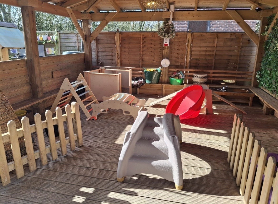 Gray plastic toddler slide sits idle, facing a red bowl-shaped rocker inside a sunlit wooden gazebo play area with low picket fences, climbing toys, storage baskets, benches, and decking.