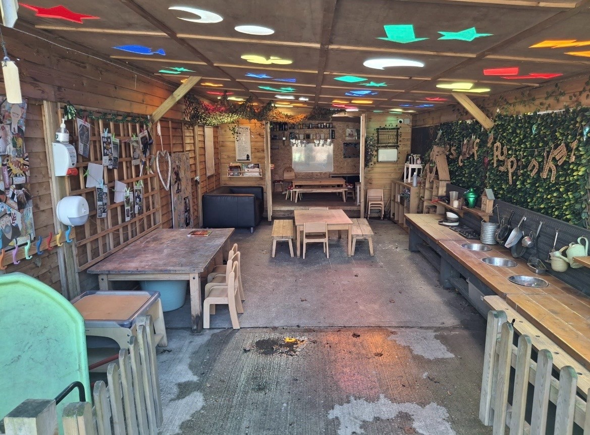 Low wooden tables and small chairs arranged for children, with a play-sink counter and craft displays; colored ceiling shapes cast multicolored light throughout a cozy wooden play shed.