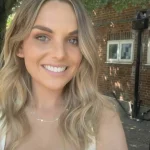 Smiling woman with long wavy blonde hair looks at the camera; wearing a delicate necklace while standing outdoors near a brick building under dappled tree shade.