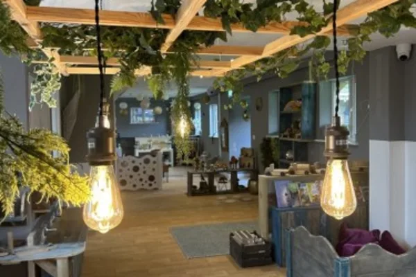 Two hanging Edison-style bulbs glow, illuminating a cozy, plant-draped interior with wooden beams overhead, rustic furniture and shelves, scattered decor, and a warm wooden floor.