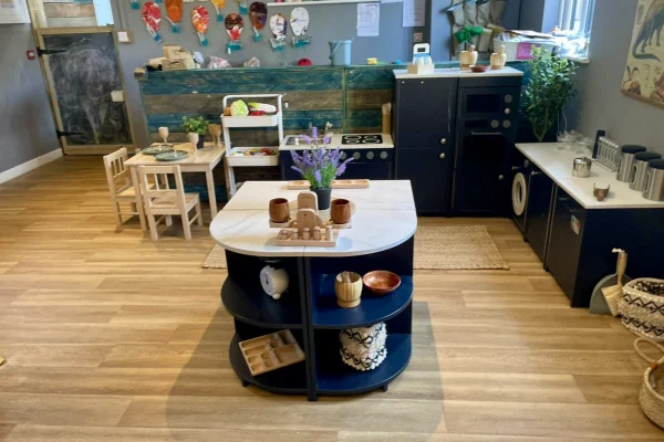 Wooden play-kitchen island holds wooden dishes, cups and a vase of purple flowers; surrounded by toy stove, sink, small table with chairs, chalkboard and baskets on a hardwood classroom floor.