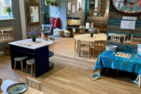 Low wooden tables and small chairs arranged for children, holding potted plants and activity materials, occupying a sunlit, tidy preschool room with wooden floors, cushions, shelves, and art displays.