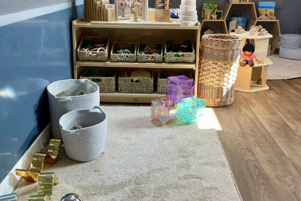 Wooden shelving with wicker baskets holds small toys; translucent iridescent cubes rest on a rug; knitted bins and a tall woven hamper flank the shelf in a sunlit playroom.