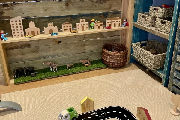 Toy road with a small green truck marked "SLOW" sits on a beige playmat, beside a wooden shelf holding miniature wooden buildings, toy animals on fake grass, and wicker storage baskets.