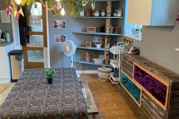 Dining table draped in patterned cloth with a potted plant; kitchen area includes a standing fan, decorative curved shelves, hanging foliage and bulbs, wood floor and glitter-paneled storage.