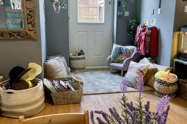 Low armchair sits angled in a cozy entry nook, surrounded by cushions, baskets of hats and magazines, a small rug, potted plants and a lavender bouquet near a back door.