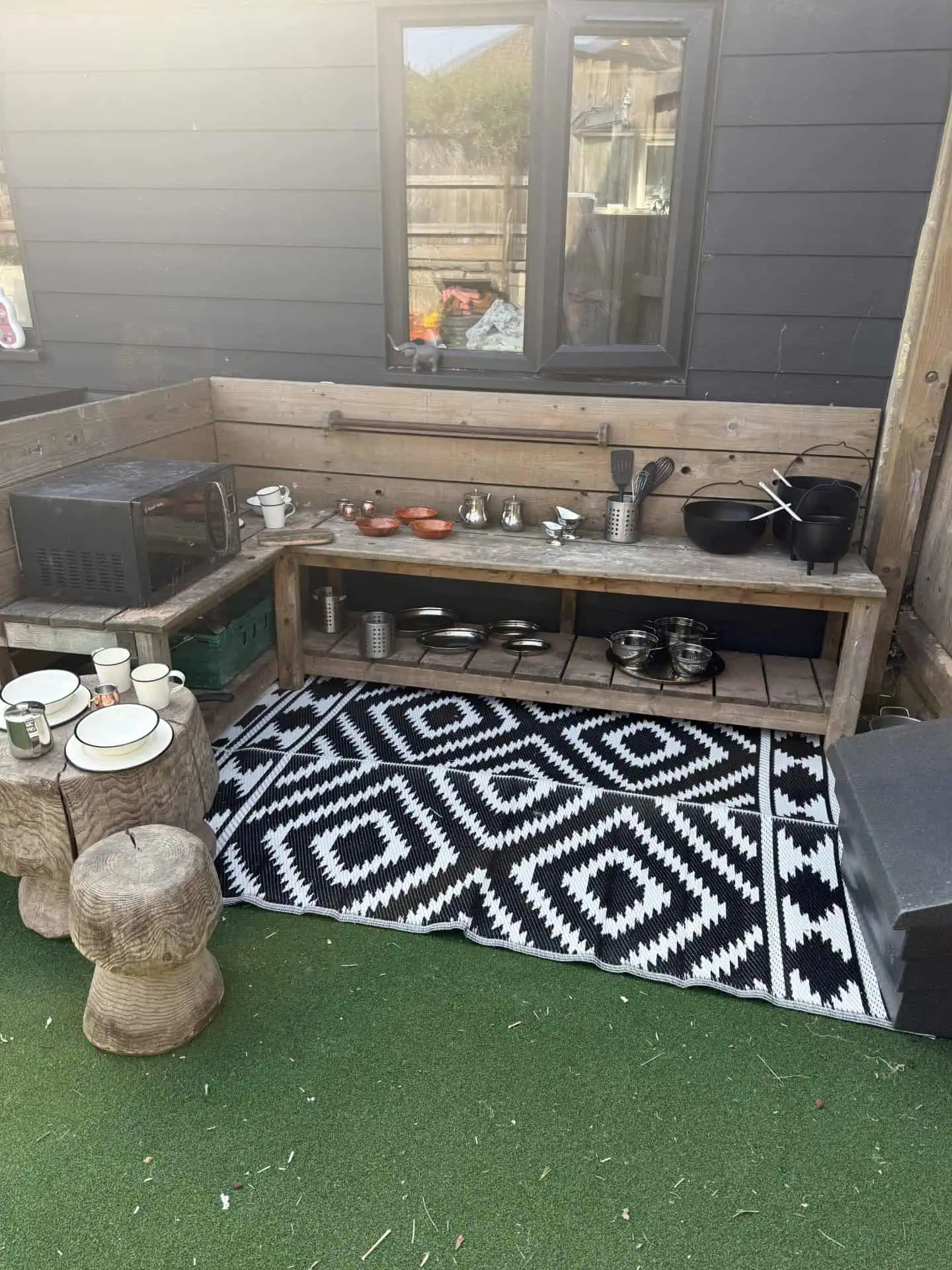 Wooden outdoor play kitchen shelf displaying pots, pans, utensils and bowls, microwave on the left, sitting on a black-and-white geometric rug over artificial grass against dark siding beneath a window.