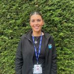 Woman standing and smiling, wearing a black jacket and blue lanyard, posed outdoors in front of a dense green hedge.