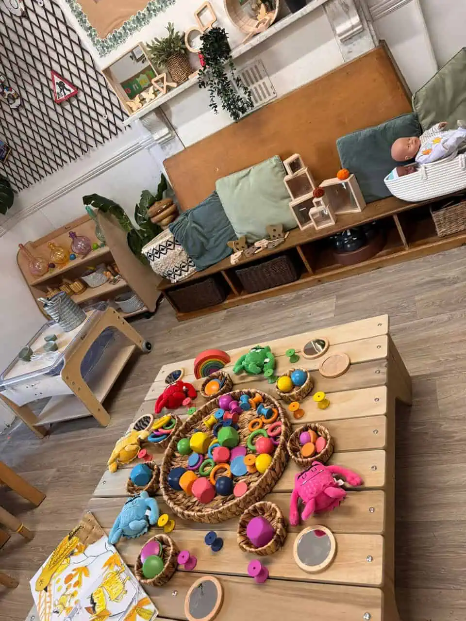 Wooden play table holds scattered colorful wooden shapes, wicker baskets and plush toys; set in a cozy children's playroom with low shelving, cushions, plants and baby doll bassinet.