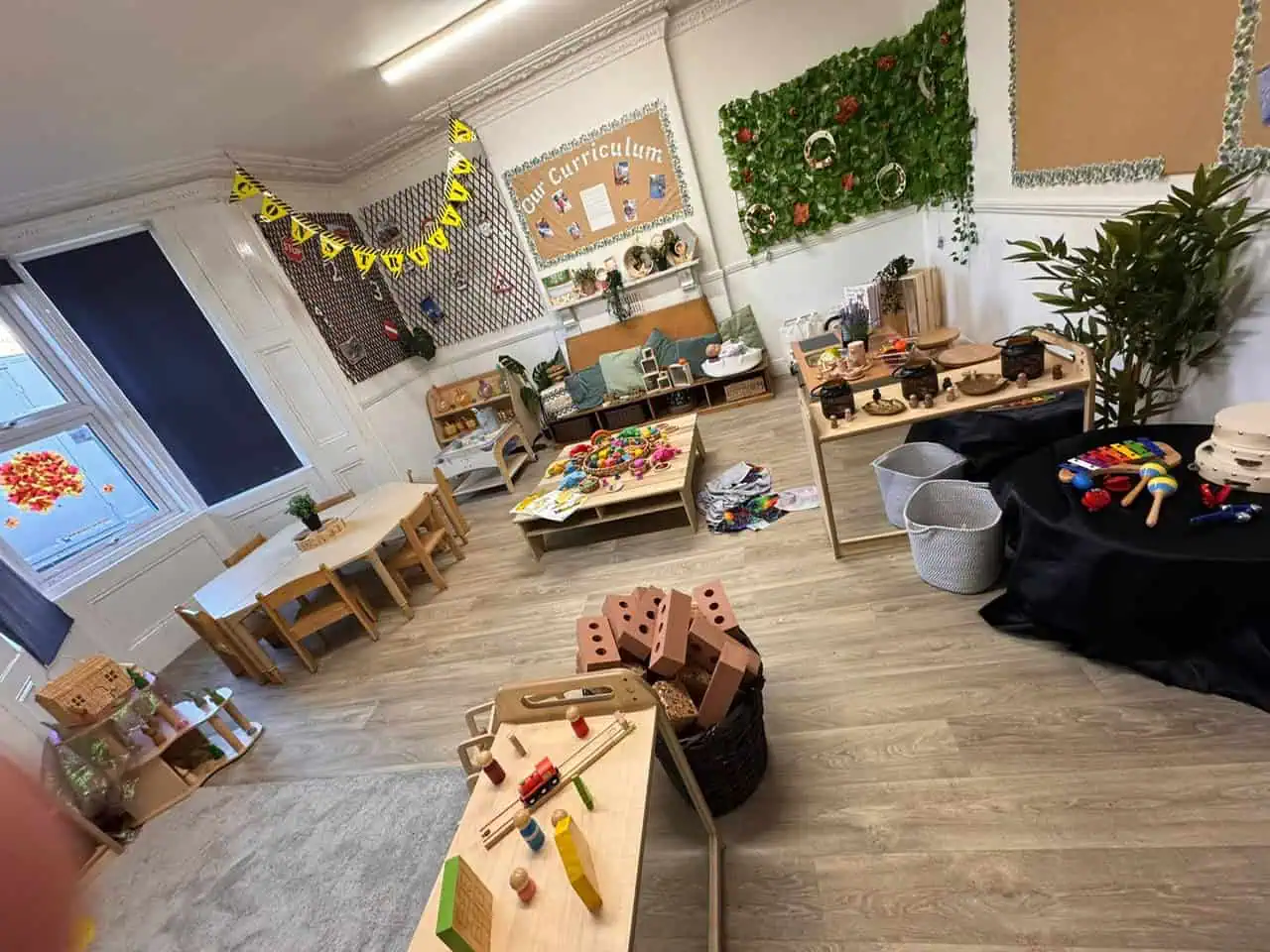 Wooden play tables and toy stations arranged for exploration; children’s toys scattered for play in a preschool classroom with tables, cushions, plants, and a bulletin board labeled "Our Curriculum".