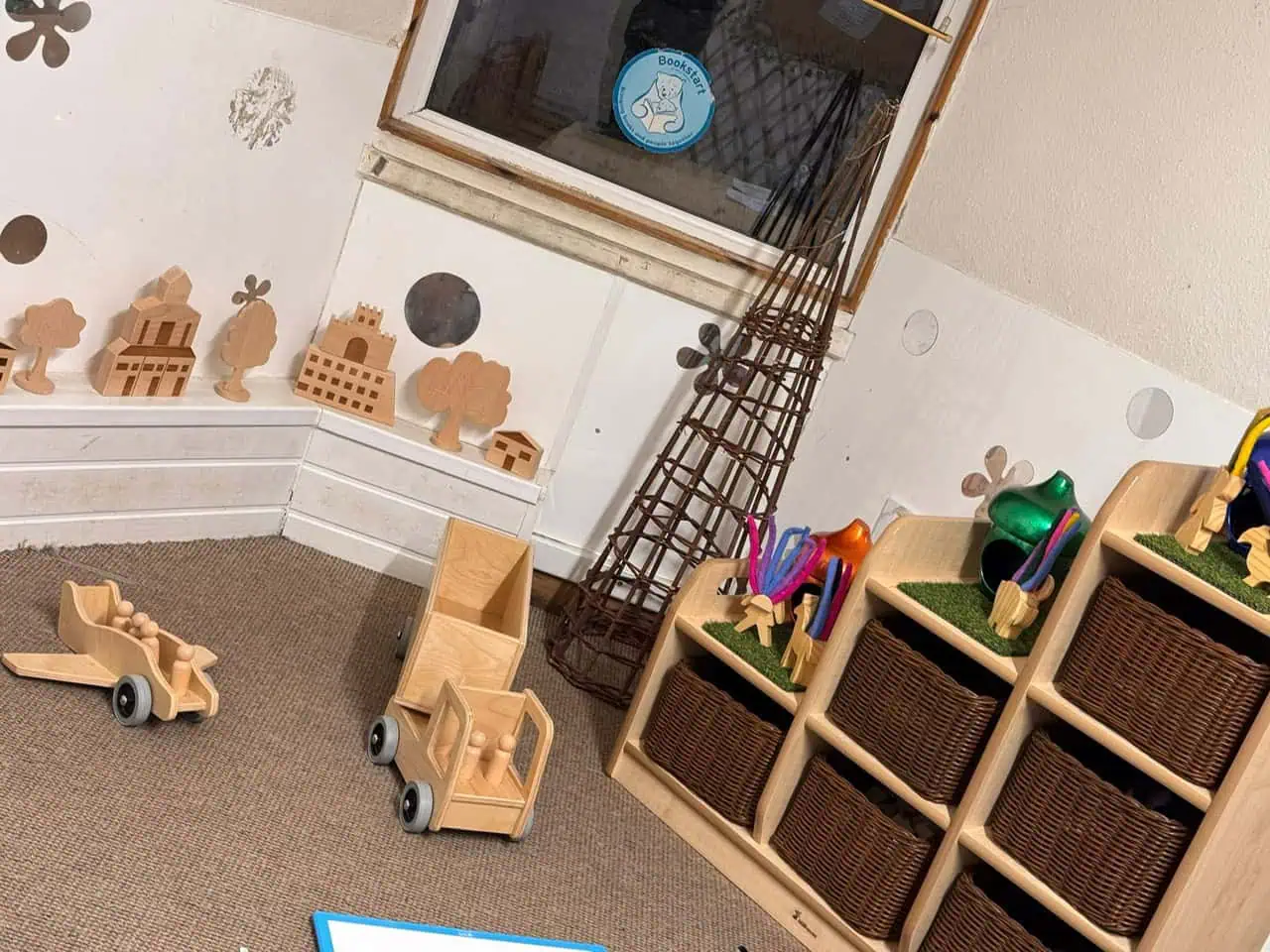 Wooden toy trucks rest on a carpeted floor near low cubby shelving holding wicker baskets and colorful craft materials, inside a small playroom beneath a window with a "Bookstart" sticker.