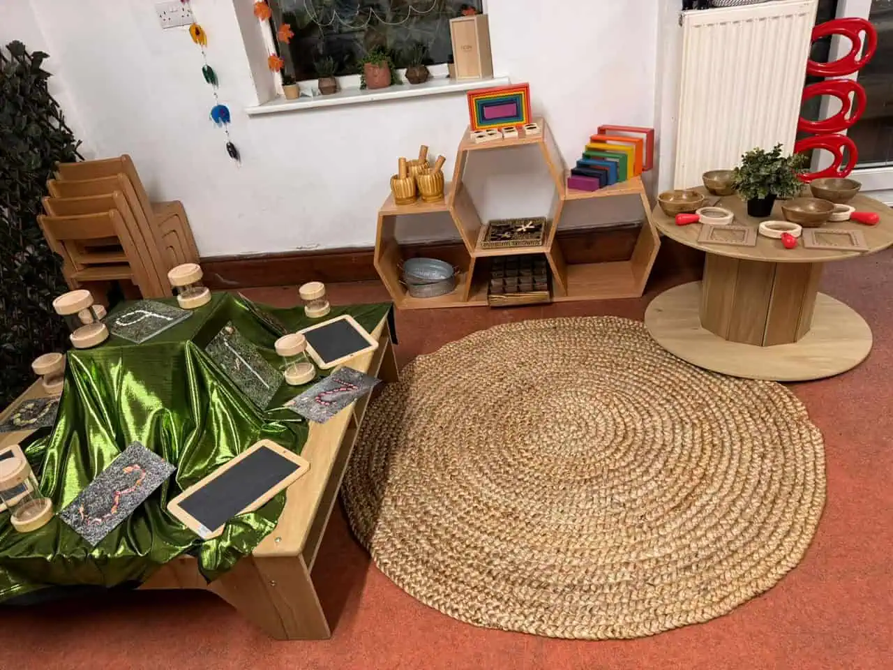 Low wooden table displays green satin, sand timers and chalkboards; hexagonal shelves hold wooden toys and rainbow blocks; braided round rug and spool table occupy carpeted classroom corner.