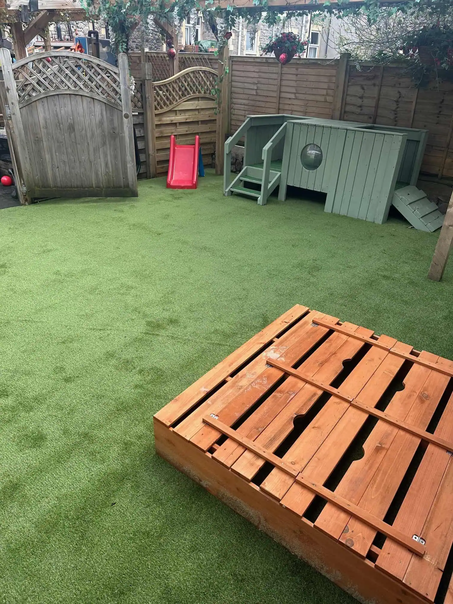 Wooden slatted sandbox sits on artificial grass; behind it, a red plastic slide and pale green wooden playhouse with steps and porthole window, enclosed by wooden fences.